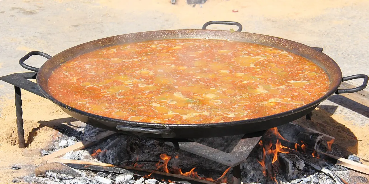 Una paellera grande hirviendo sobre unas patas de hierro encima de un fuego de leña, con el caldo aún burbujeando antes de que se seque el arroz