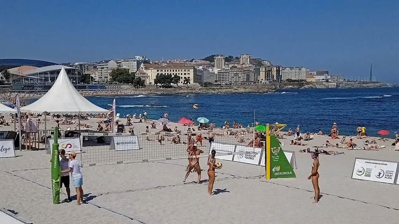 Beachvolleyballers op een zandveld tijdens een toernooi aan zee, met een witte tent en een stad op de achtergrond