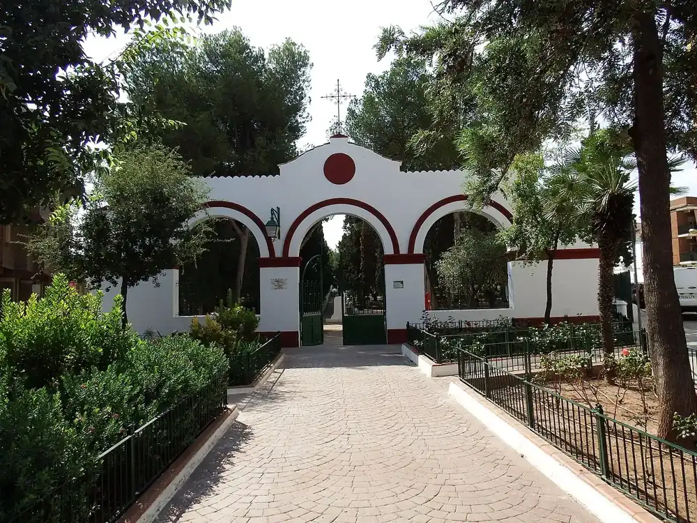 A garden square with a stone statue and a white arched gateway among pine trees in Bétera