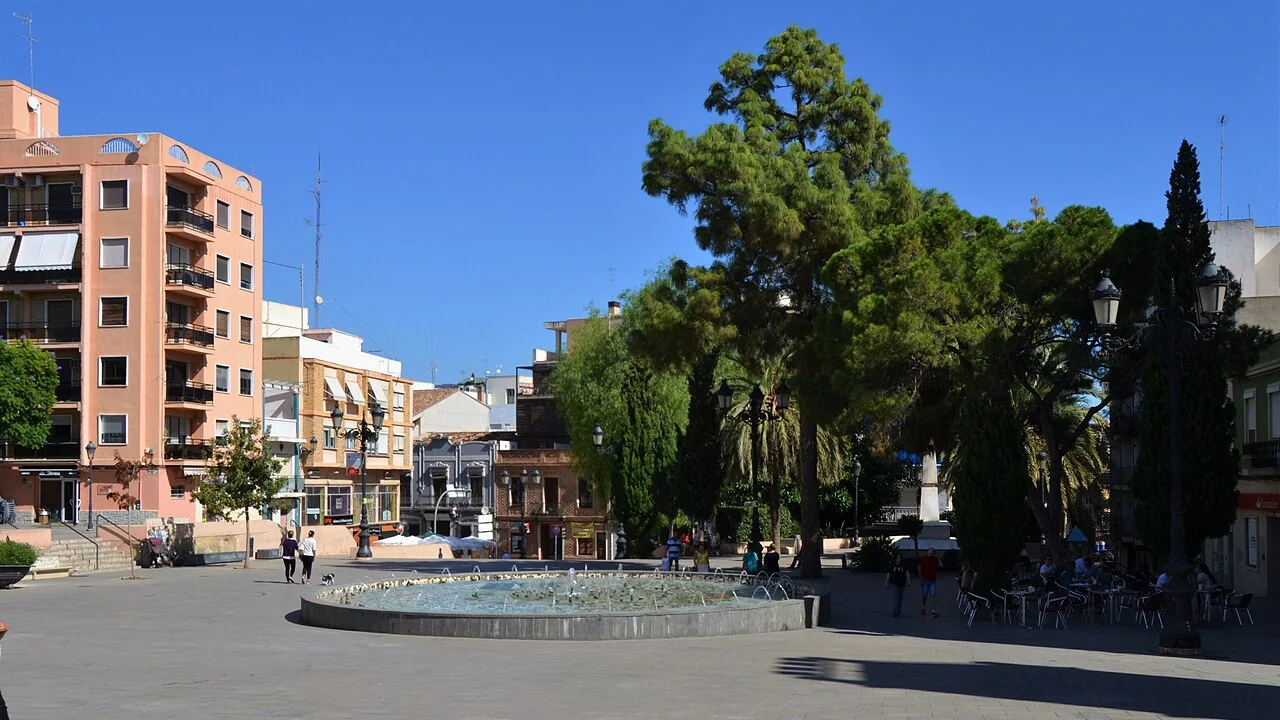 La plaza de Emilio Castelar de Burjassot, una plaza empedrada con una fuente redonda, pinos altos y terrazas de café al sol