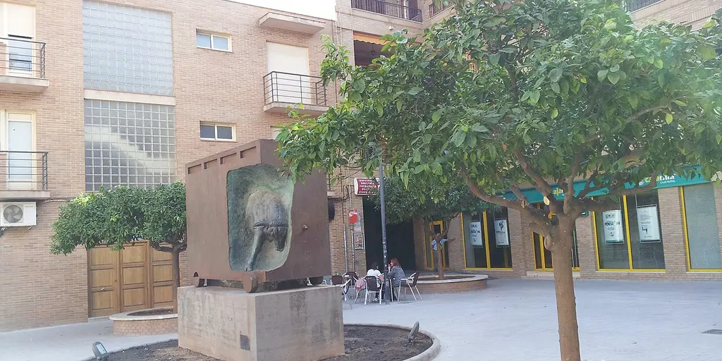 A small paved square with a stone monument and young trees in Pucol