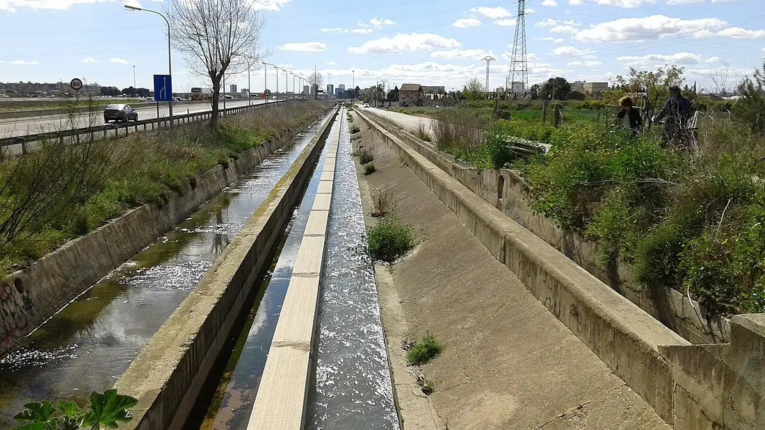 Un canal d'irrigation à l'eau vive le long d'une route importante, avec de hauts immeubles de Valencia au loin