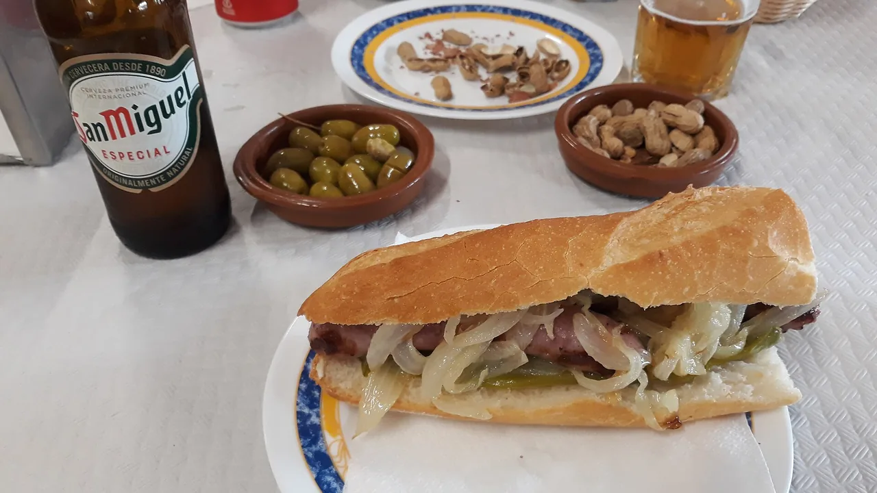 A baguette sandwich with grilled sausage and onions on a plate, beside bowls of olives and peanuts and a bottle of beer