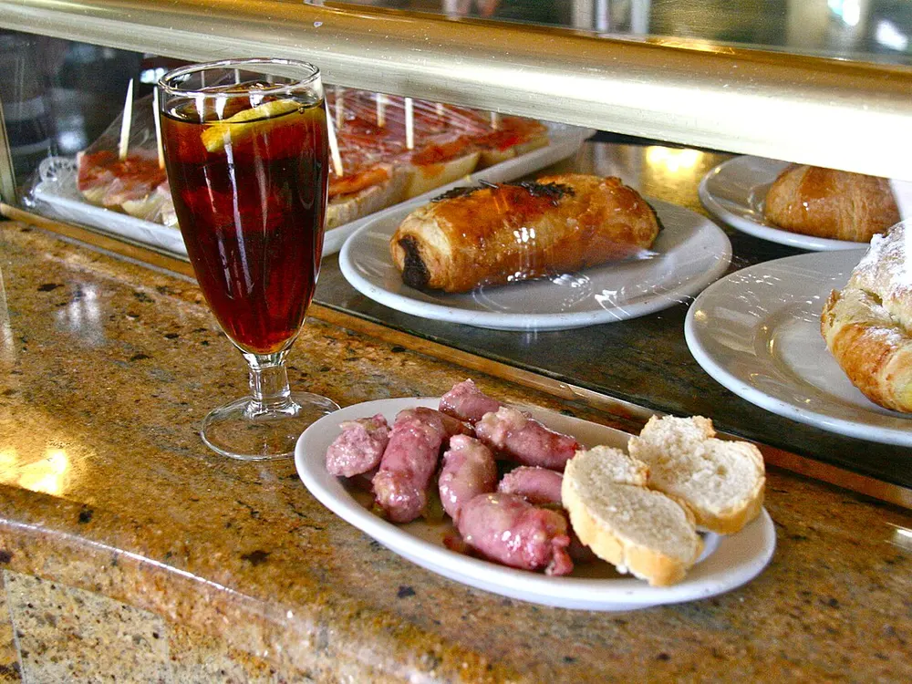 Una copa de vermut rojo con rodaja de naranja y hielo junto a platos de tapas en la barra de un bar español