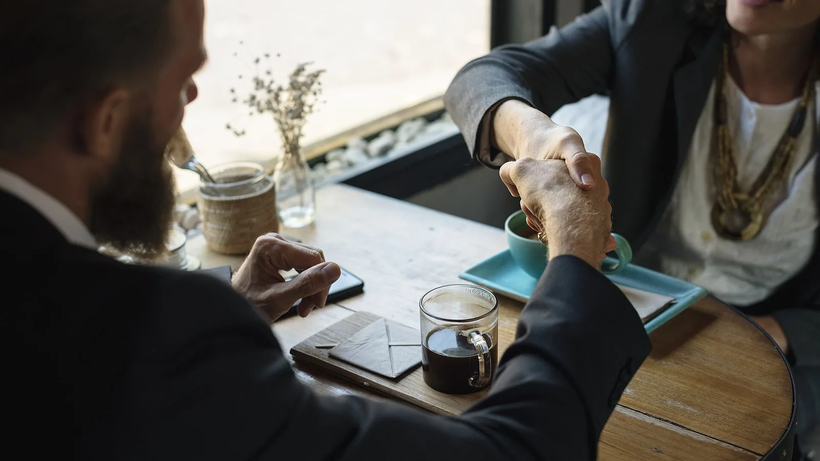 Two people shaking hands across a small table with coffee cups between them, one wearing a dark blazer, the other in a white top with layered necklaces