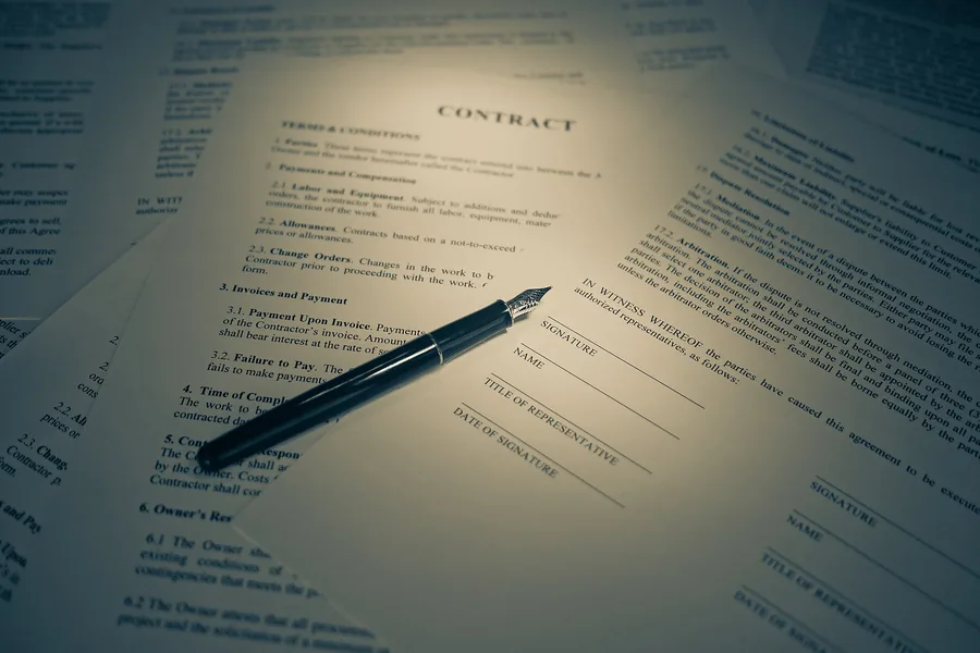 A pen resting on a printed contract's signature page, warm desk lamp light, other papers fanned out around it