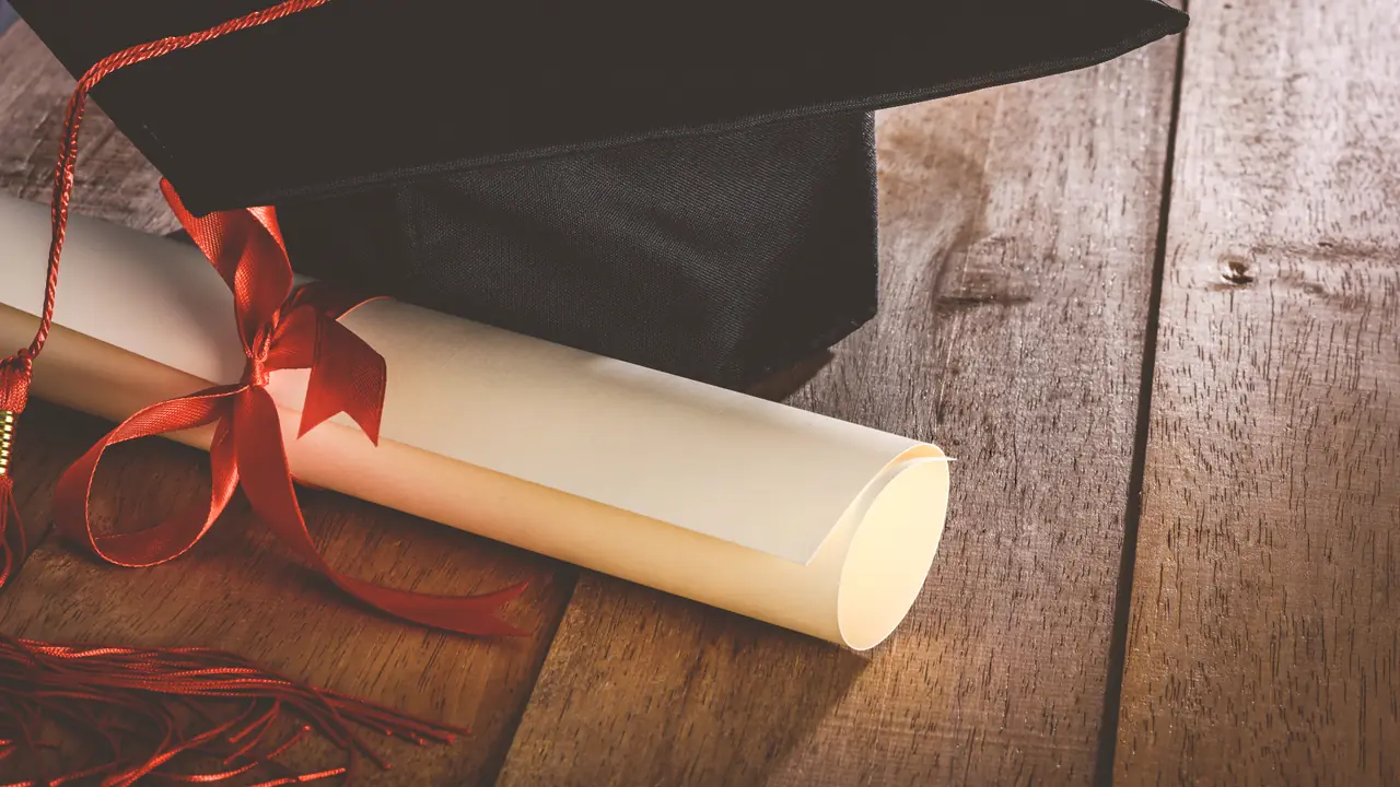 Een afstudeerhoed en een opgerold diploma met een rood lint op een houten bureau